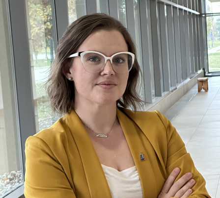 Eva Yelle, Director at Uplift, wearing white-framed glasses and a mustard blazer, standing in a sunlit hallway